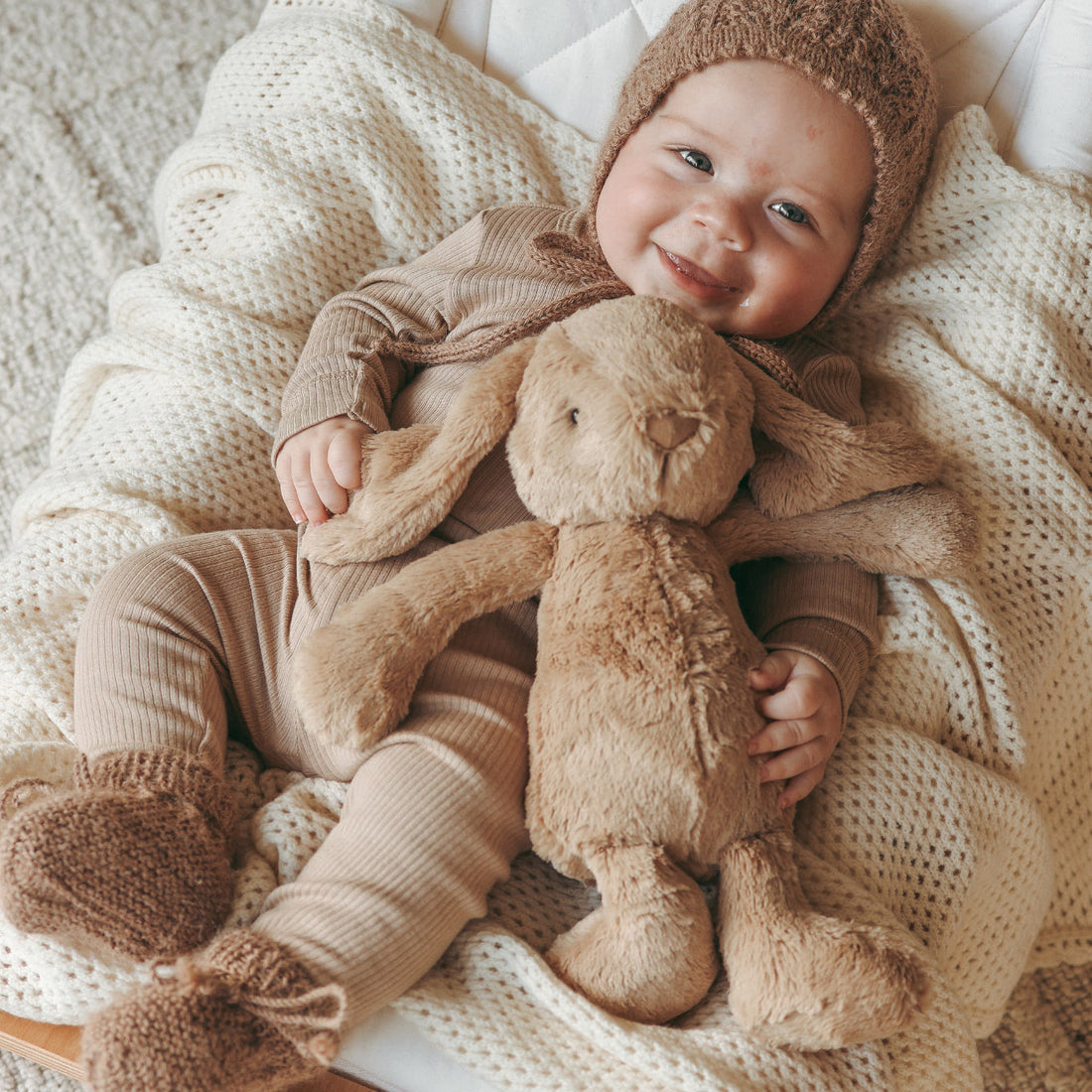 Baby in brown outfit and hat, holding a stuffed bunny, laying on a white blanket in a wooden cradle.