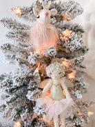 A close-up of a flocked Christmas tree decorated with a stuffed deer ornament with fluffy pink fur and a fairy doll with a white halo, golden wings, and a pink tutu.