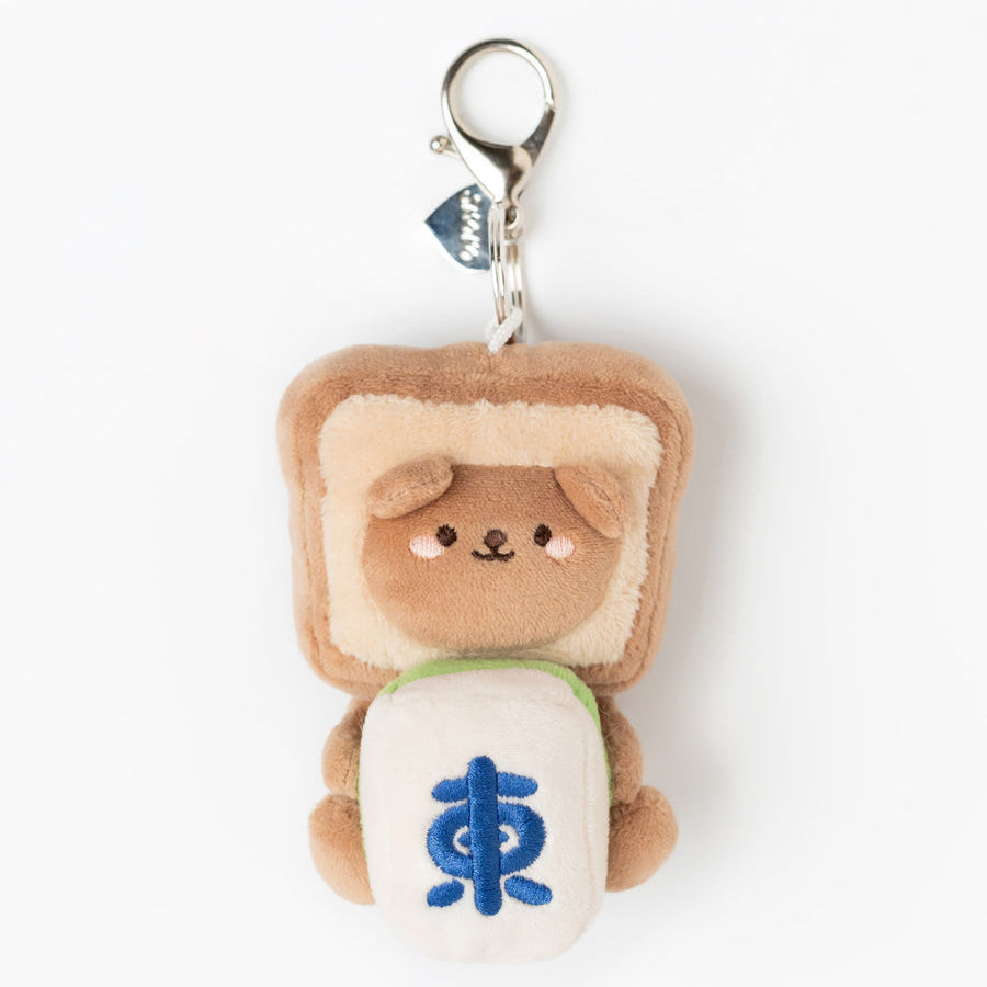 A plush keychain charm shaped like a cute brown bear wearing a tan bread slice hat and holding a white bun with a blue symbol, attached to a silver metal clasp on a white background.