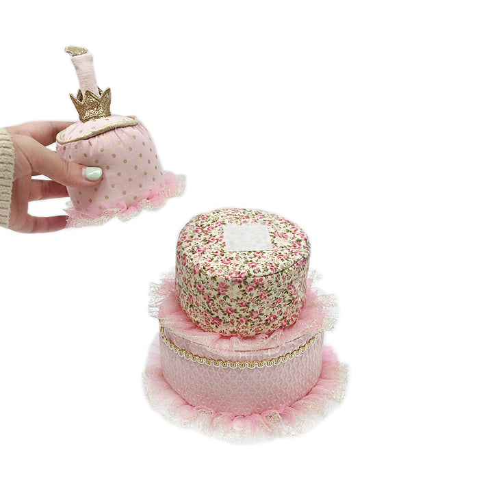 A pink fabric-covered 2-tier cake with lace trim and a hand holding a matching bell-shaped doll with a crown.