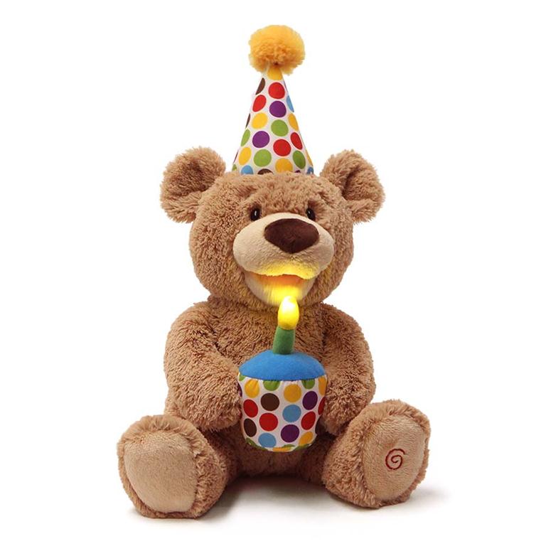 Brown teddy bear wearing a polka dot party hat and holding a lit birthday candle cupcake.