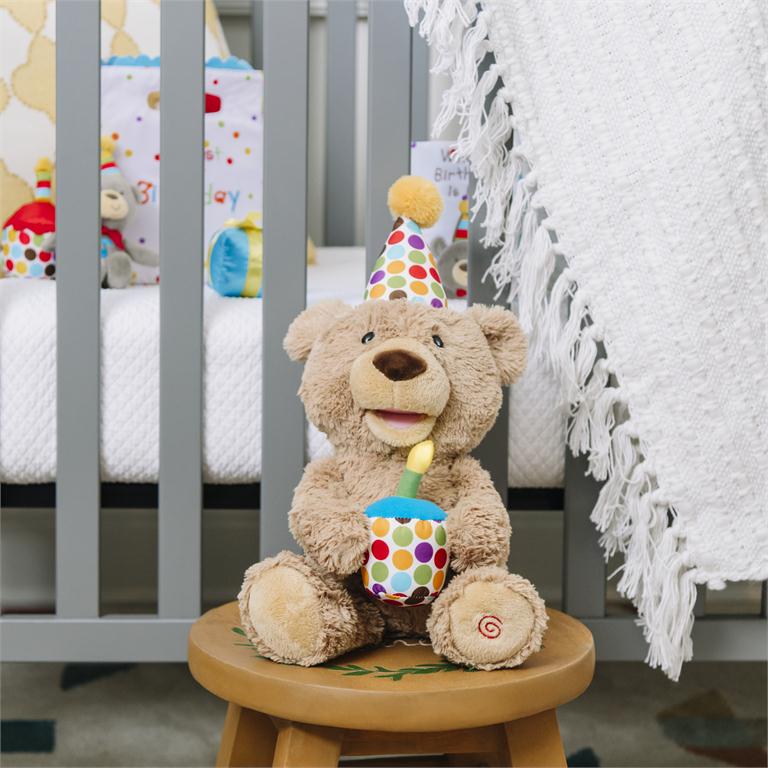A stuffed teddy bear wearing a polka-dot party hat and holding a candle sits on a wooden stool in front of a gray crib.