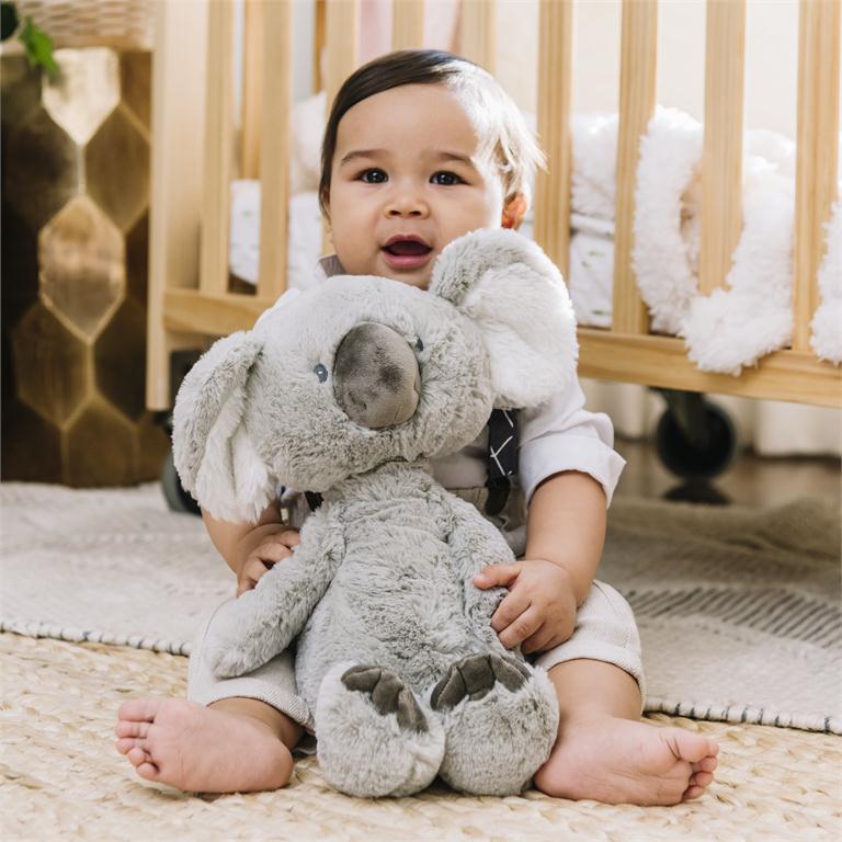 Baby holding gray plush koala bear, wearing cream clothing and suspenders, seated on a woven rug.