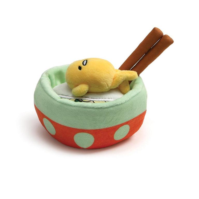 A plush yellow toy shaped like an egg yolk is inside a soft, circular bowl with a green top, an orange bottom with green polka dots, and two brown chopsticks sticking out of the back.