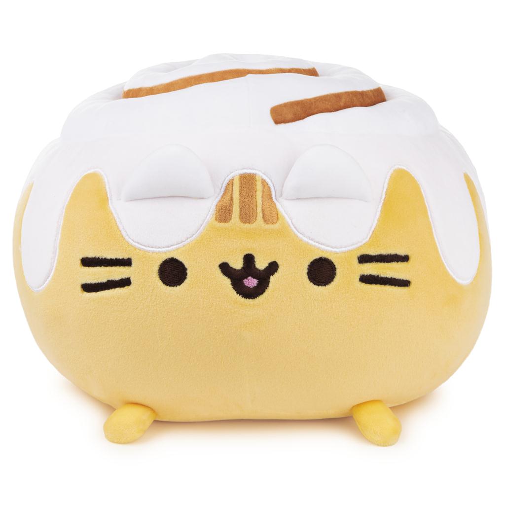 Pusheen cat plush resembling a cinnamon roll. The plush is light yellow, with white icing details and brown swirl on top. It has small legs and a smiling face.