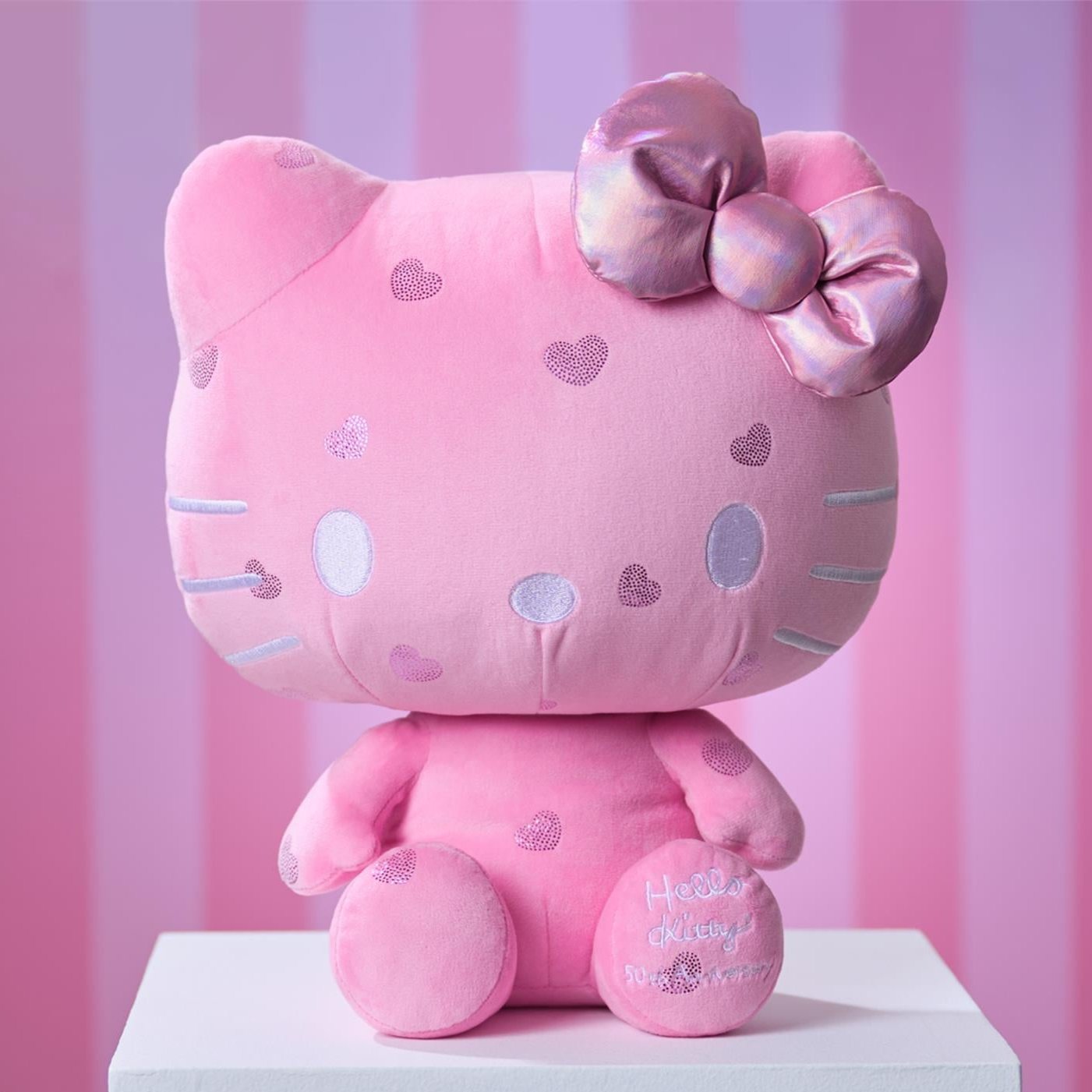 Pink plush Hello Kitty doll with shiny bow and heart accents, sitting on a white surface against a pink striped background.