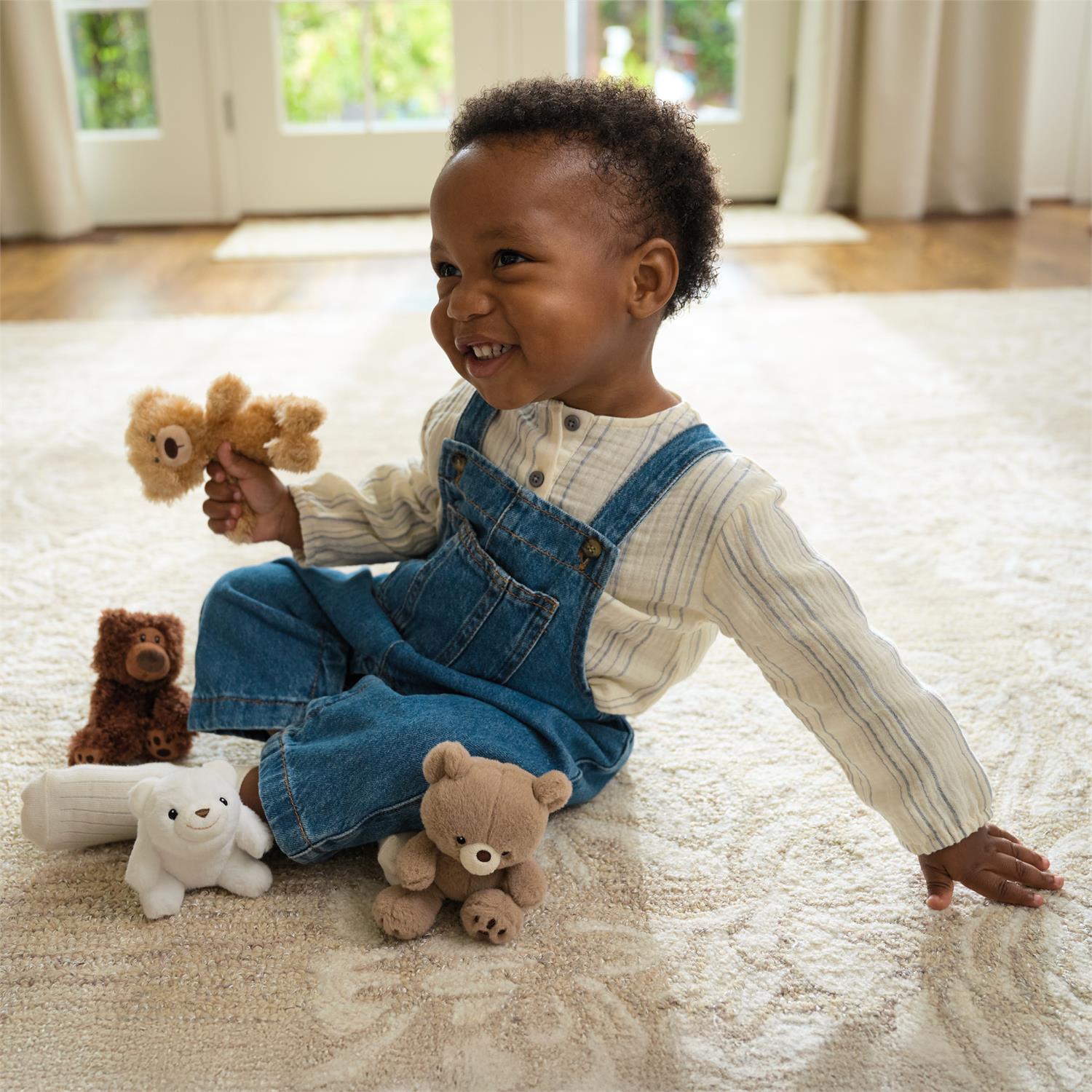 Smiling child in denim overalls sitting on a rug surrounded by teddy bears.