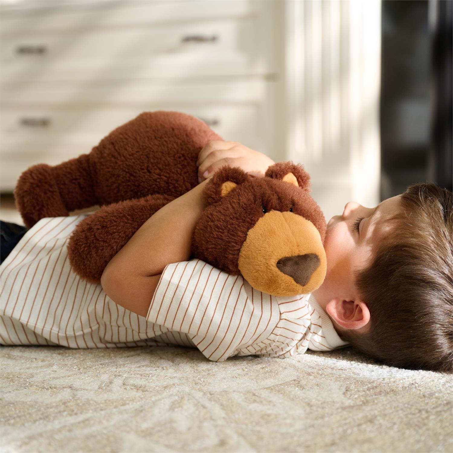 Boy lying on rug, hugging brown teddy bear, wearing striped shirt.