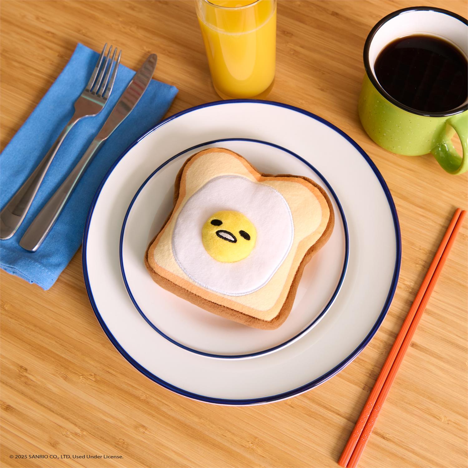 Overhead shot of an egg-on-toast plush toy on a plate, with a fork, knife, napkin, orange juice, coffee, and chopsticks on a wooden table.