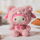 Pink plush My Melody toy in a fuzzy bear costume, seated indoors with baked goods in the background.