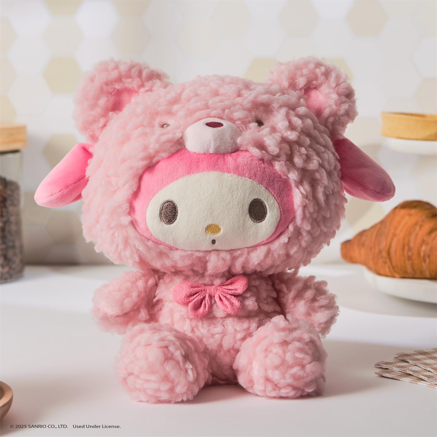Pink plush My Melody toy in a fuzzy bear costume, seated indoors with baked goods in the background.