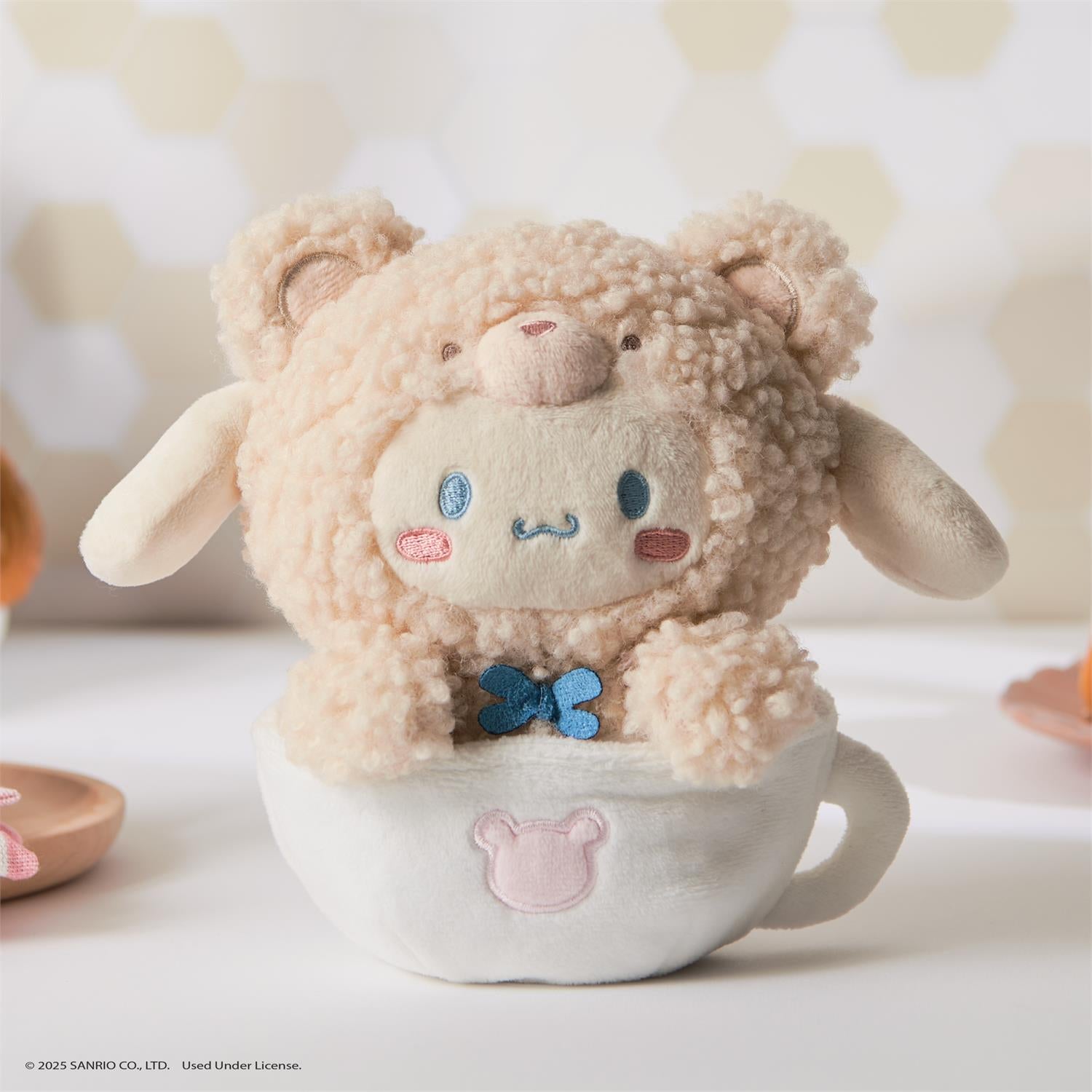 Stuffed animal in a teacup. The stuffed animal is beige with a fluffy bear-like outer layer and a white face with blue eyes and a pink nose. It has a blue bow around its neck. The teacup is white with a pink bear-shaped patch on the front.