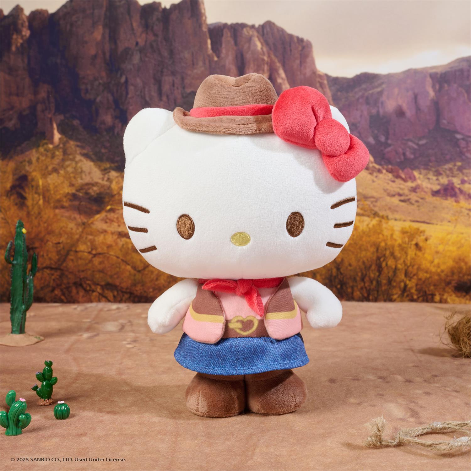 Hello Kitty plush toy dressed as a cowboy in a desert setting with cacti and mountains.