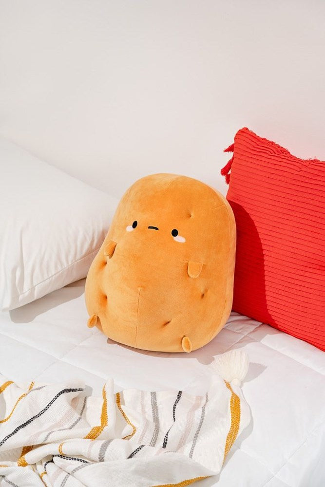 Stuffed plush potato toy with face, on a bed, next to white pillow and red pillow with fringe. Striped blanket in foreground.