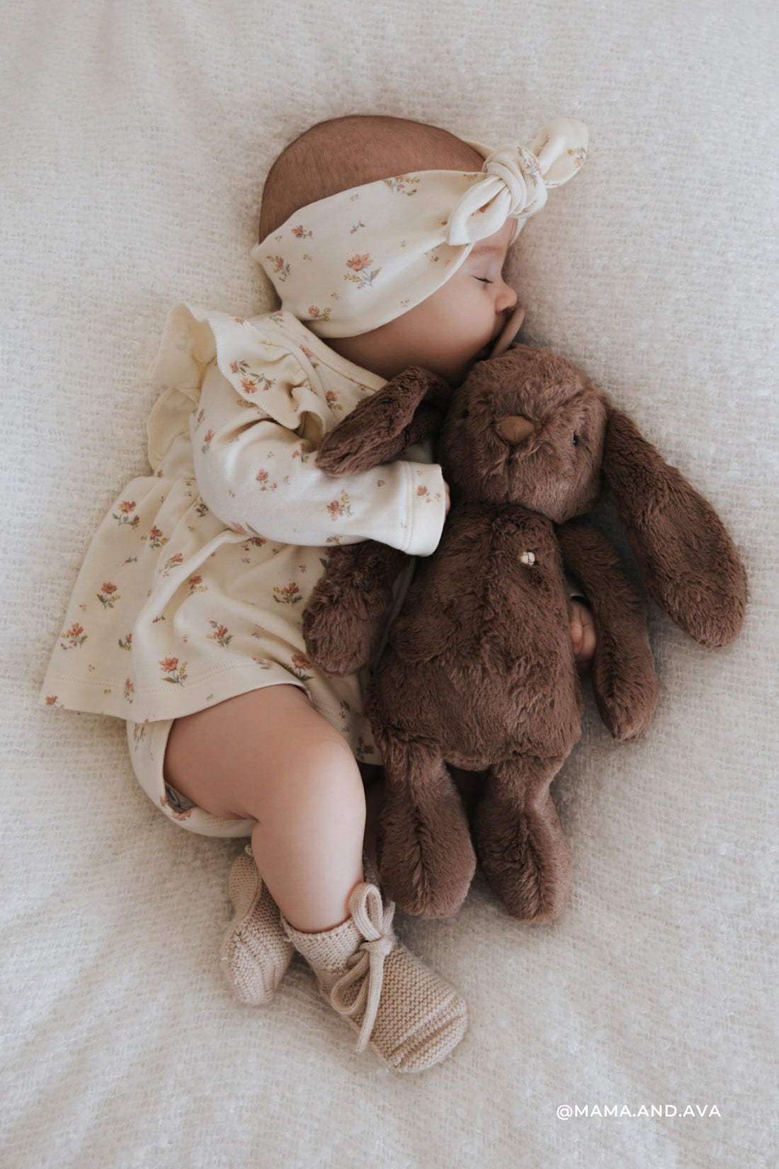 Sleeping baby with floral headband, dress, booties, and brown plush bunny.