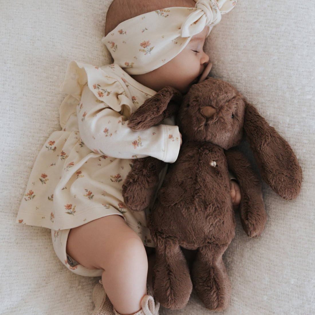 Sleeping baby with floral headband, dress, booties, and brown plush bunny.