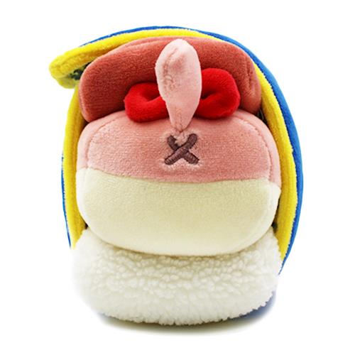 Plush toy of a character with pink face and red eyes, a brown 'nose', and a stitched 'X' on its face. It's surrounded by yellow and blue trim, resting on a fluffy white base.
