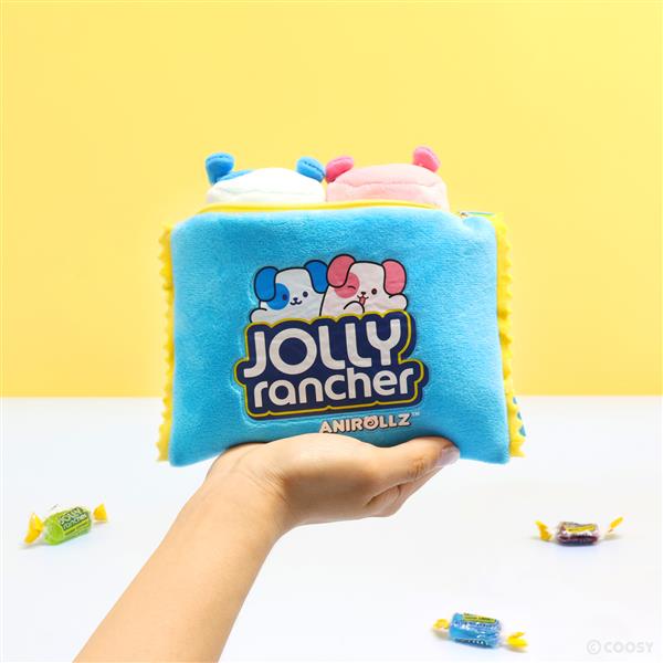 A hand holding a blue plush pouch with a "Jolly Rancher Annirollz" design, with Jolly Rancher candies scattered on a white surface in front of a yellow background.