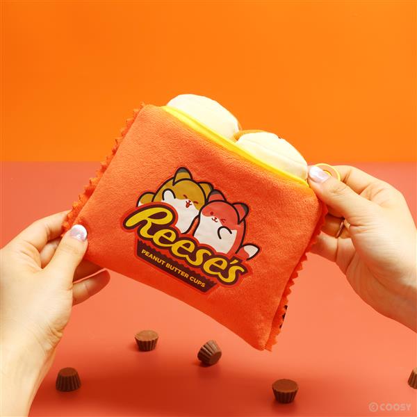 A person holds an orange zippered Reese's Peanut Butter Cups-themed plush bag that contains two peanut butter cup-shaped plush toys, with several real miniature peanut butter cups scattered on the orange surface below.
