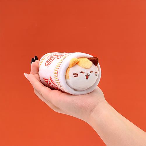 A plush cat in a ramen noodle cup held in a person's hand.