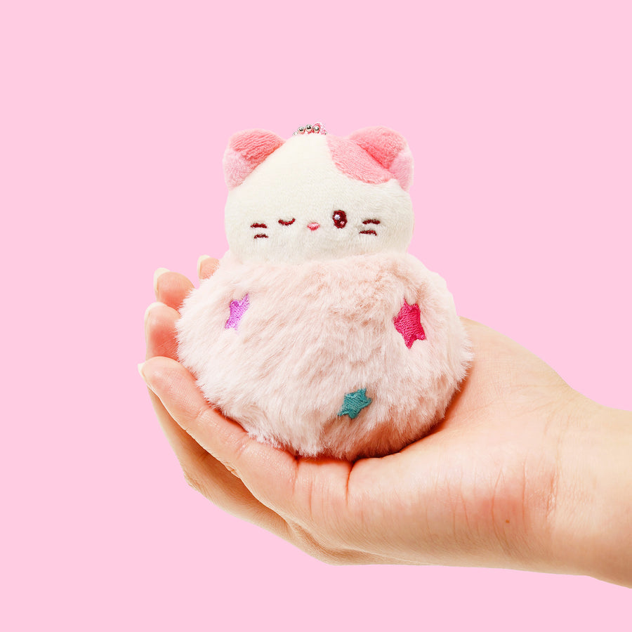 Plush toy resembling a cat with pink cheeks held in a hand against a pink background