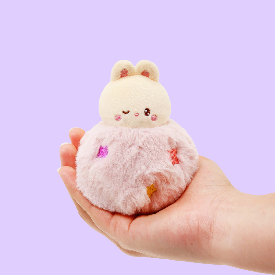 Plush toy resembling a cat in a dress held in a hand against a purple background