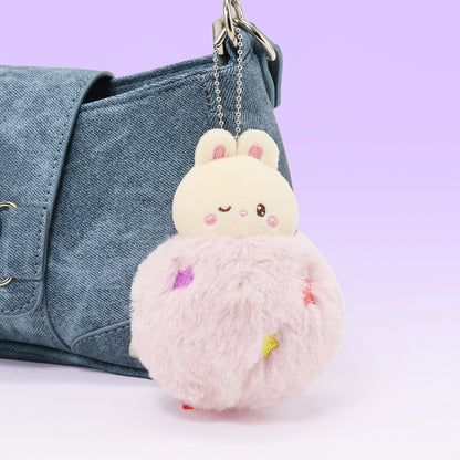Plush keychain shaped like a bear in a pink dress hanging from a denim bag on a light purple background