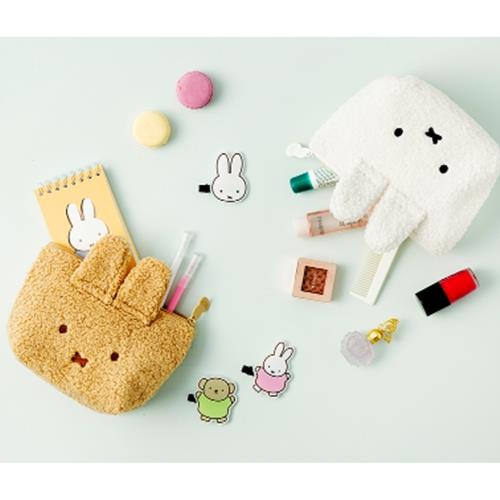 Overhead shot of a beige furry pouch with bunny ears, yellow notebook, pastel macarons, and a white furry pouch with bunny ears scattered with makeup, clips, and other accessories on a light green surface.