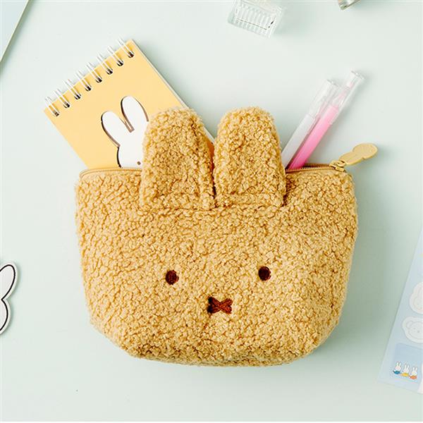 Close-up of a fluffy, tan bunny-shaped pouch with a yellow notebook and pens inside, on a light blue surface.