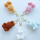 Five fluffy bunny keychains in tan, pink, brown, white, and light blue with gold-tone hardware.
