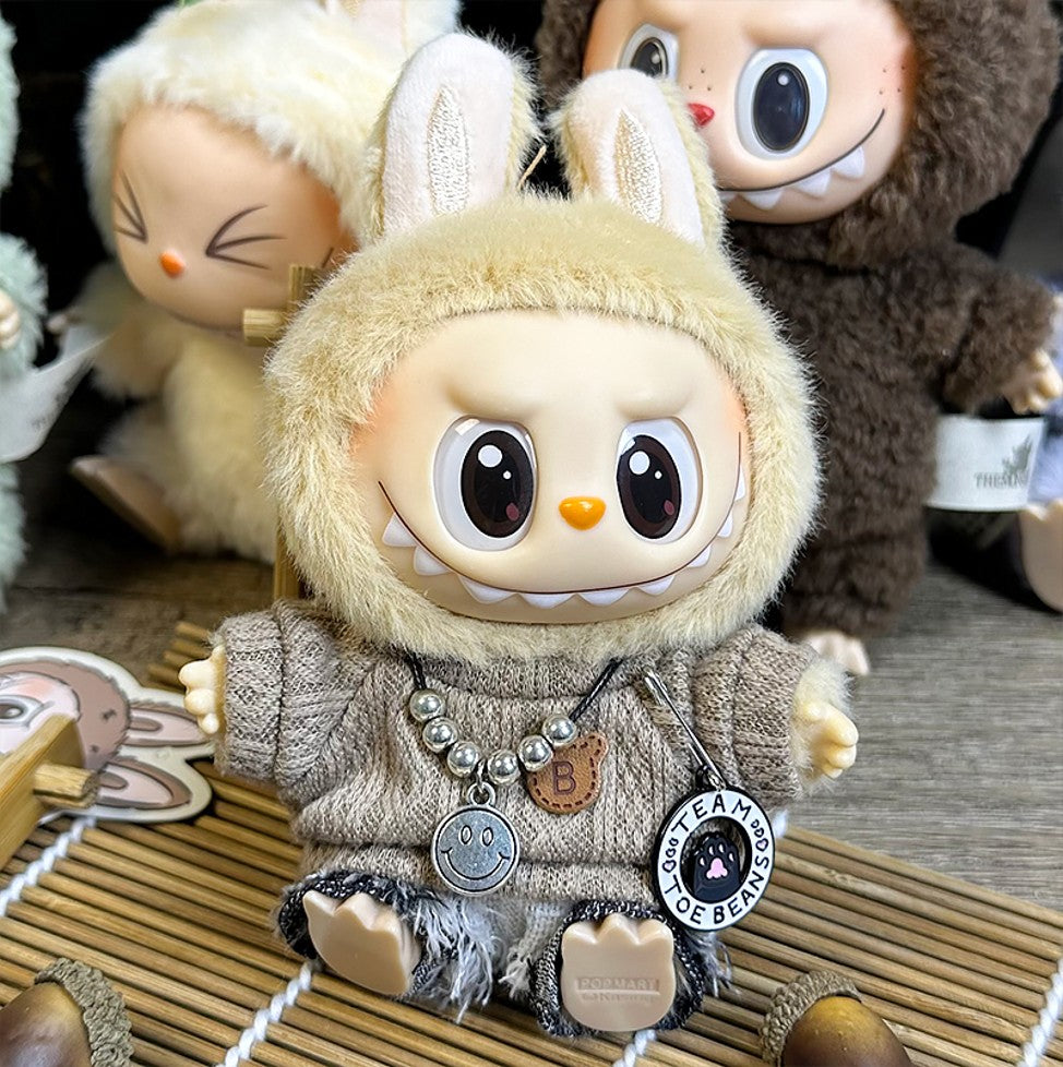 Close-up of a toy doll dressed in a beige sweater and bunny hood, featuring a necklace with charms, sitting on a bamboo mat.
