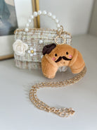 Small plush dog keychain with a mustache and bow tie on a gold chain, placed on a light surface with a decorative bag in the background.