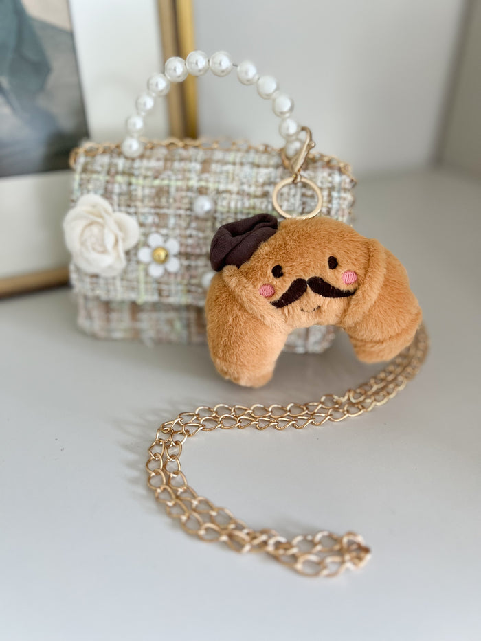 Small plush dog keychain with a mustache and bow tie on a gold chain, placed on a light surface with a decorative bag in the background.