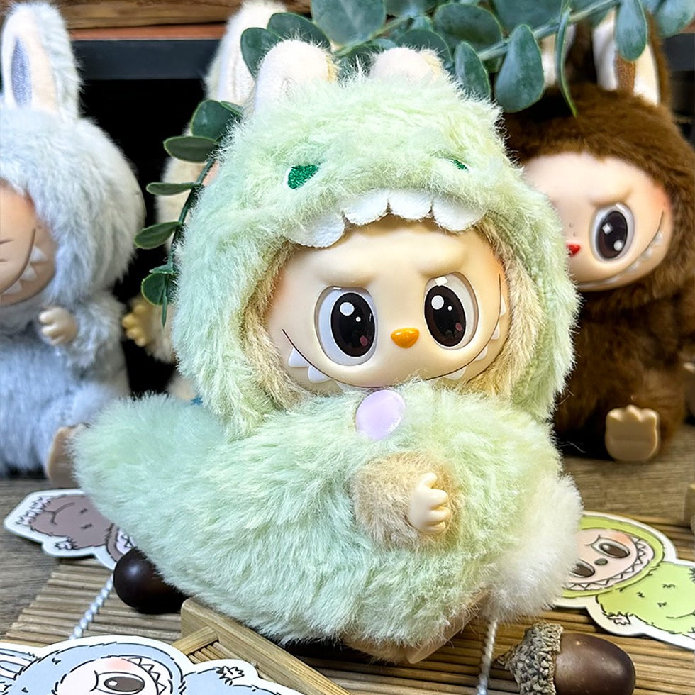Close-up of three plush dolls. The doll in the center wears a fuzzy, green dinosaur costume. The dolls beside it wear fuzzy gray and brown bunny costumes. The dolls are on a wooden surface with stickers and acorns.