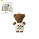 Crocheted brown teddy bear wearing white overalls with colorful bird designs.