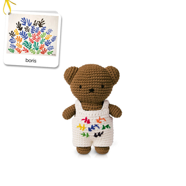Crocheted brown teddy bear wearing white overalls with colorful bird designs.