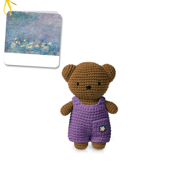 Crocheted brown teddy bear wearing purple overalls with a flower detail. A painting is in the top left.
