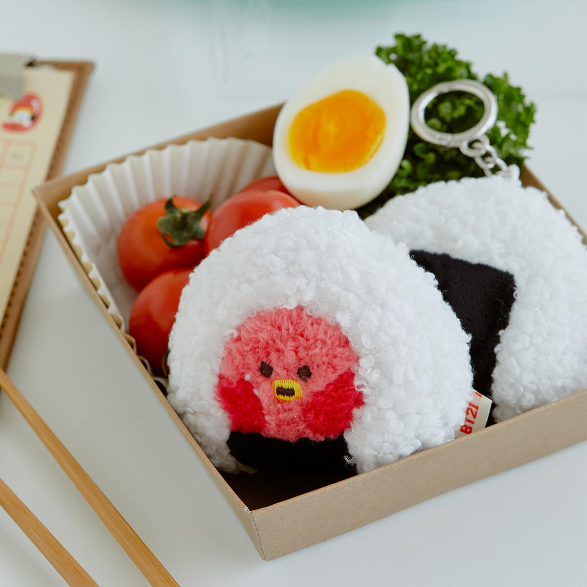 A bento box containing plush onigiri rice balls with character faces, tomatoes, a sliced egg, and a green sprig with a key ring.