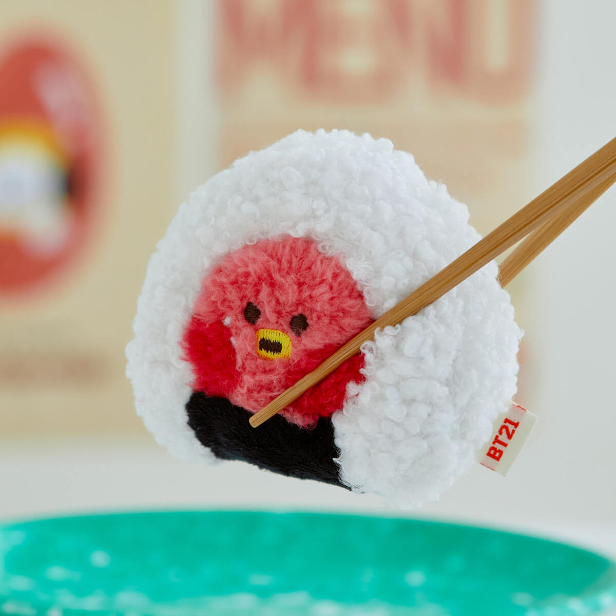 Plush sushi toy being held by chopsticks. The toy is white and red with a black accent at the bottom, and has a yellow beak. The "rice" is a textured white material. A "BT21" tag is visible. A green plate is in the foreground.