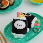 Two sushi-shaped plush keychains rest on a green speckled plate, one featuring a white, fuzzy character face.