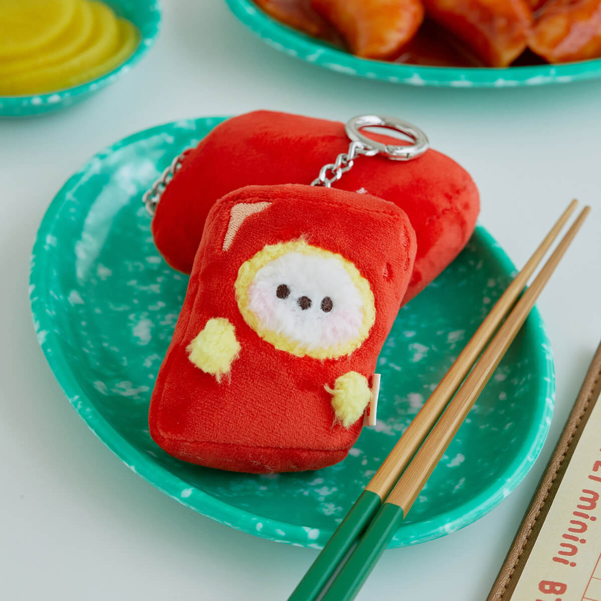 Red tteokbokki plush keychain on a turquoise plate with chopsticks.