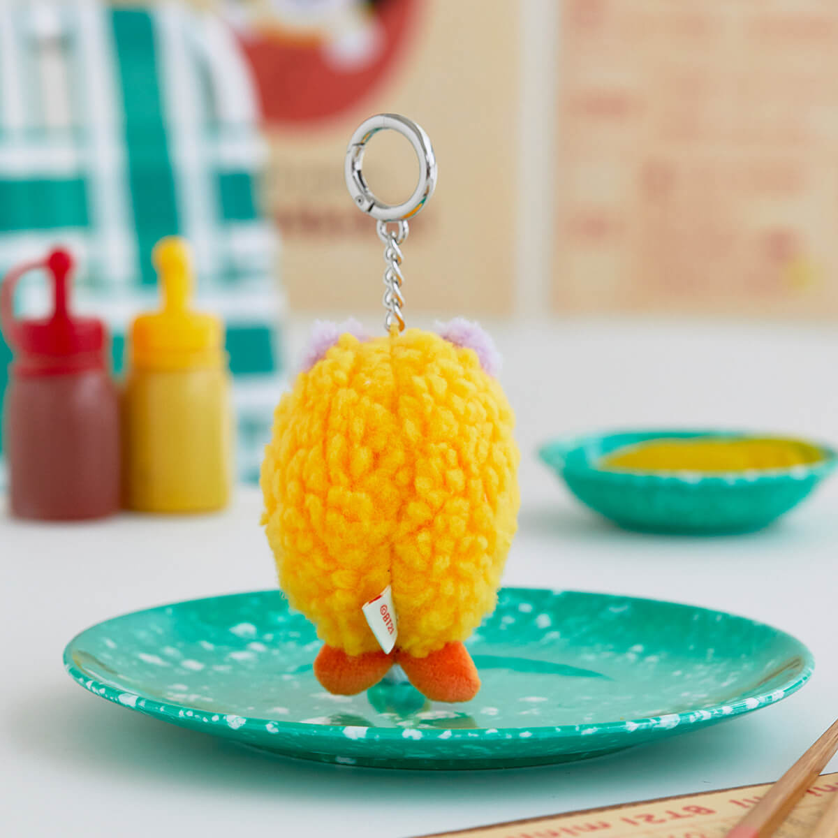 Rear view of a yellow, fluffy character keychain with orange feet and purple ears, hanging over a turquoise and white speckled plate.