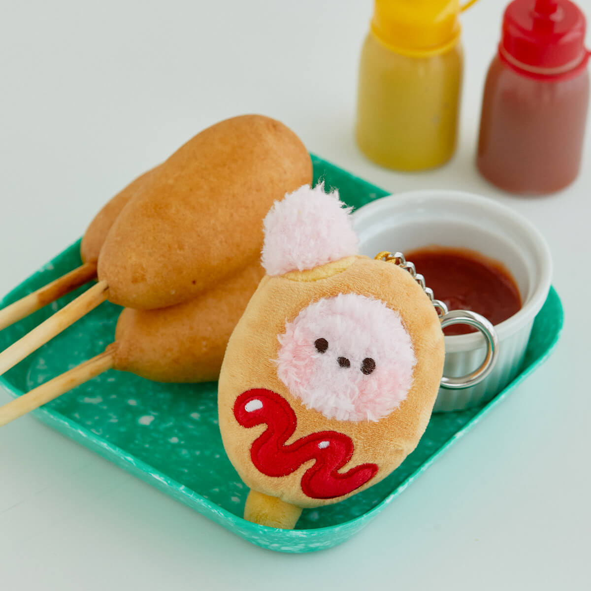 Three corn dogs, a corn dog plush keychain, ketchup, and mustard on a green tray.