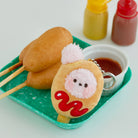 Three corn dogs, a corn dog plush keychain, ketchup, and mustard on a green tray.