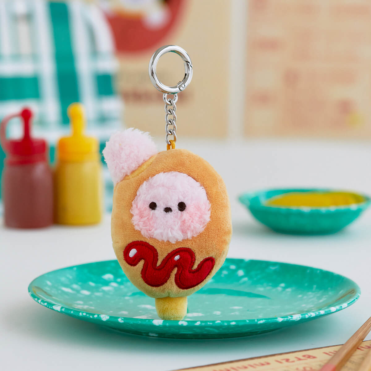 Close-up of a plush keychain shaped like a corndog with a pink poodle face and red ketchup, resting on a teal speckled plate.
