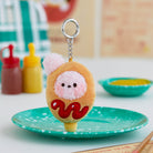 Close-up of a plush keychain shaped like a corndog with a pink poodle face and red ketchup, resting on a teal speckled plate.