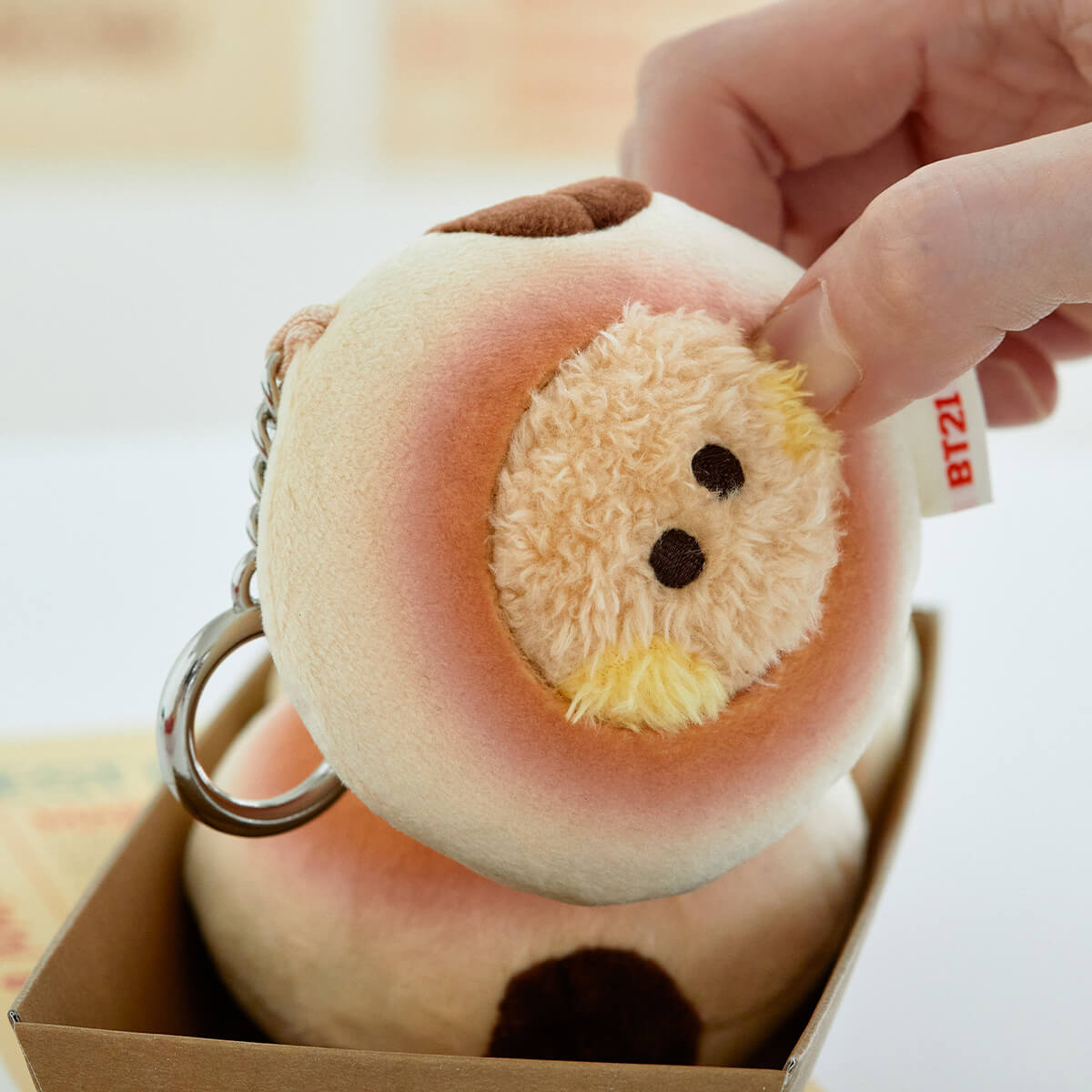 Close-up of a peach-shaped plush keychain with a furry face, held by a hand, inside a brown box.