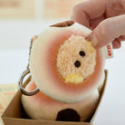 Close-up of a peach-shaped plush keychain with a furry face, held by a hand, inside a brown box.