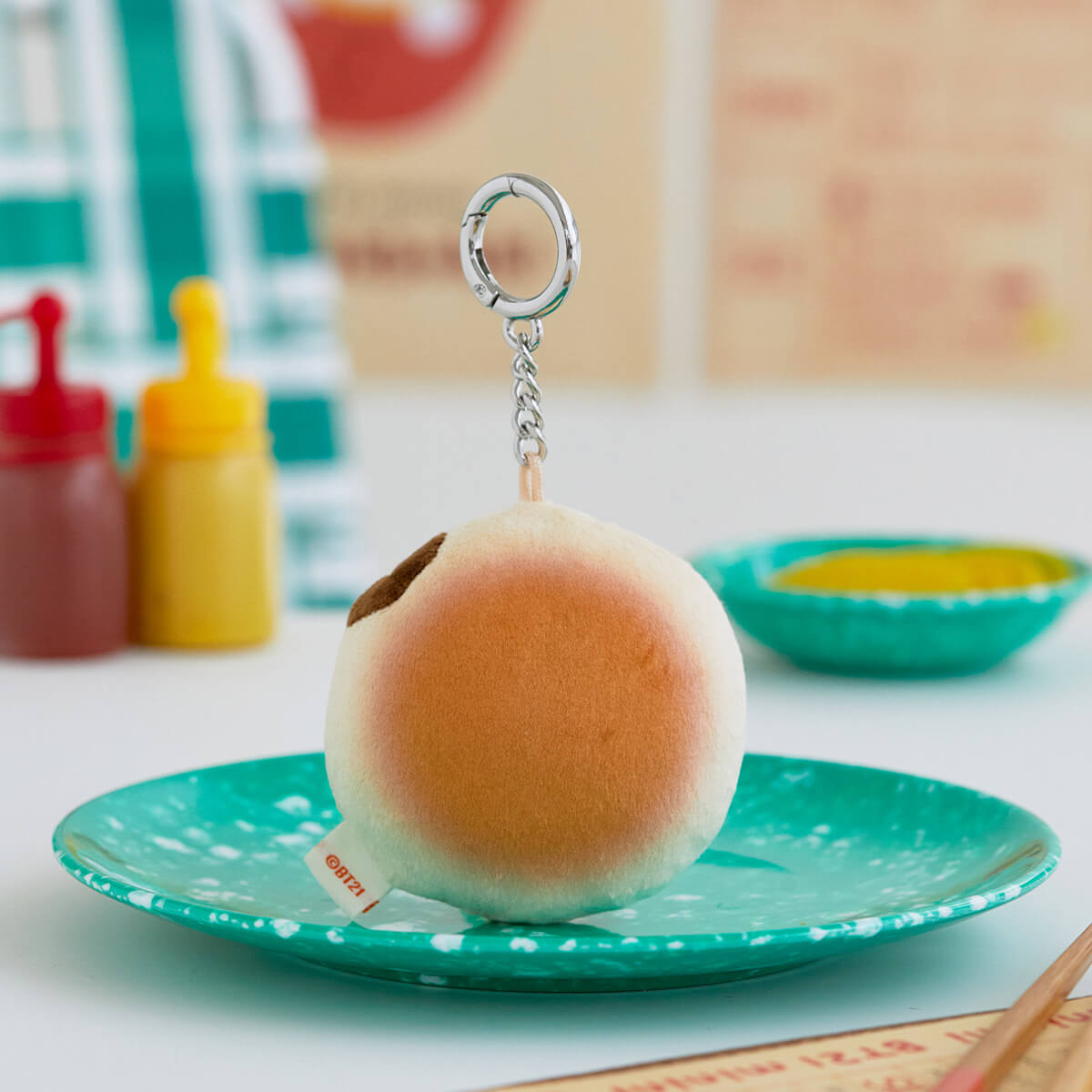 Plush BT21 bun keychain on a turquoise speckled plate, with bottles of condiments in the background.