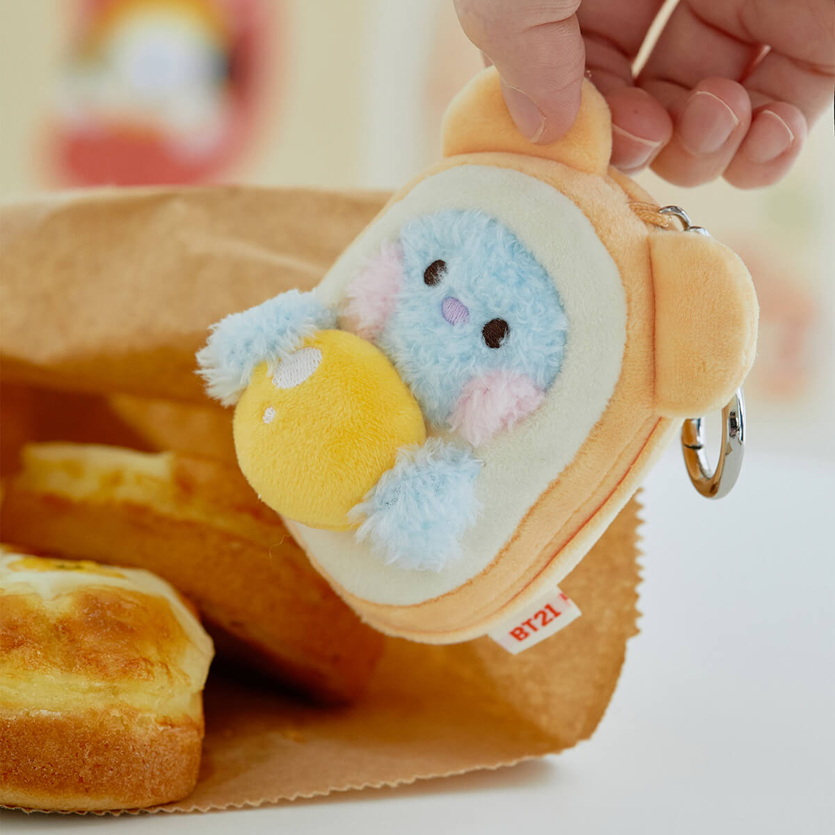 Hand holding a BT21 plush coin purse with a keyring, inside a brown paper bag with baked goods.